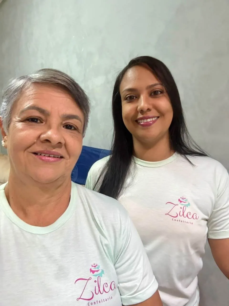 Zilma e Carol, da Zilca Confeitaria, sorrindo lado a lado com o uniforme da marca
